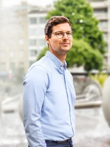 dreiviertel Portrait des Rechtsanwalt Jakob Kreutzer. Ein Mann im weißen Hemd und Brille und achsblonden Haaren, steht vor dem Brunnen am Kölner Ebertplatz