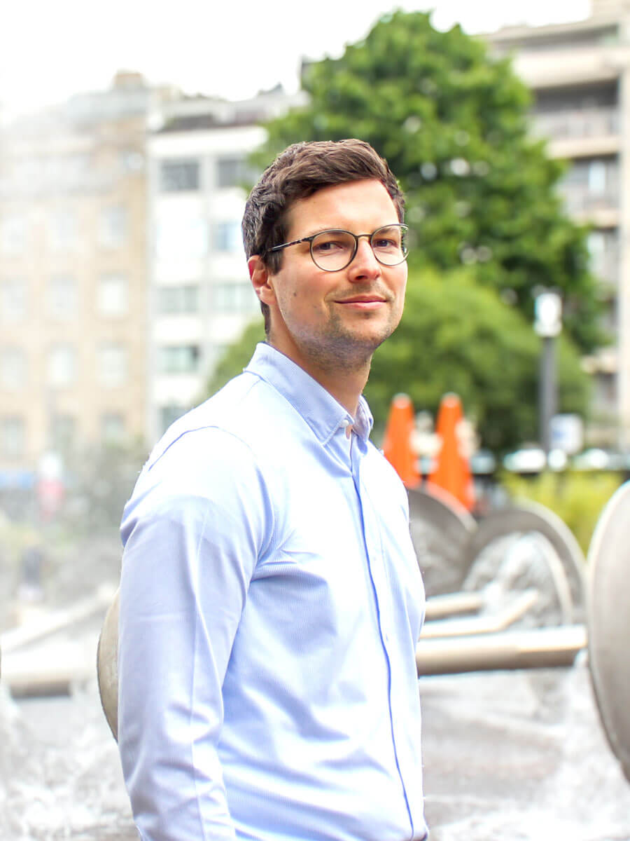 dreiviertel Portrait des Rechtsanwalt Jakob Heering. Ein Mann im weißen Hemd und Brille und achsblonden Haaren, steht vor dem Brunnen am Kölner Ebertplatz