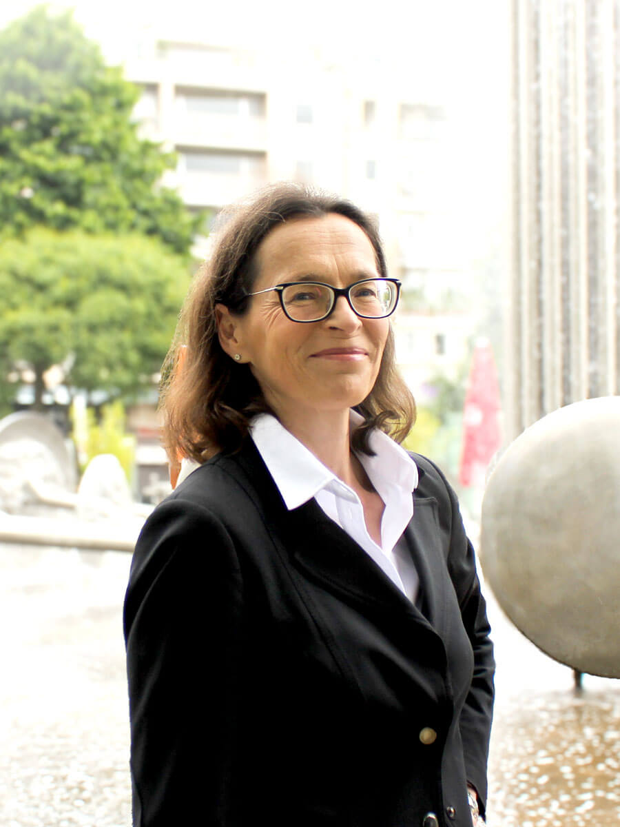 Portrait der Rechtsanwältin Sibylle Krenzel. Eine Frau mittleren Alters mit halblangen brünetten Haare und Brille. Sie steht vor dem Brunnen am Ebertplatz in Köln